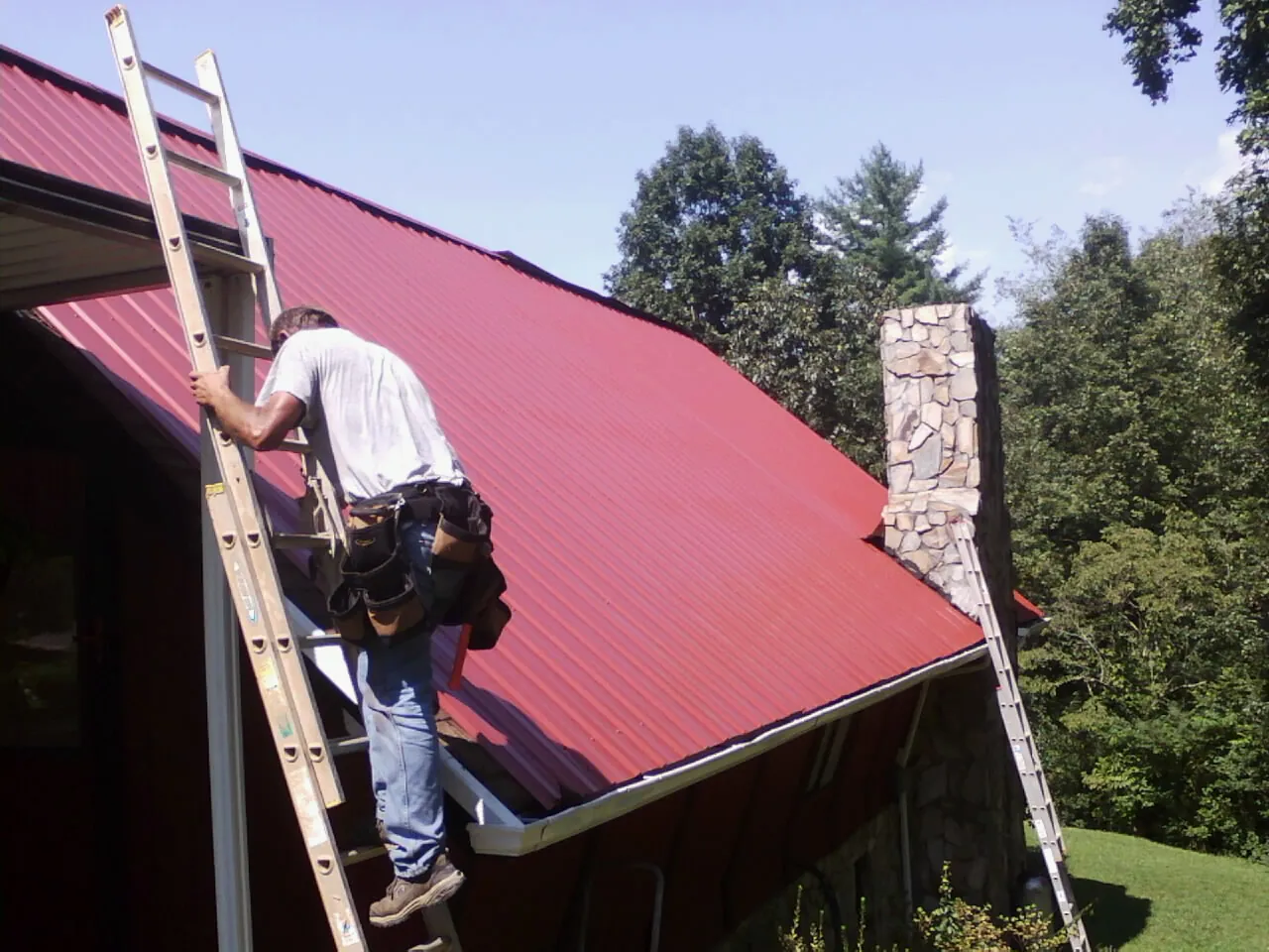 Roofer on a ladder beside a red metal roof and stone chimney — Soffit & Fascia Repair by Roofing Brothers in Mount Clemens